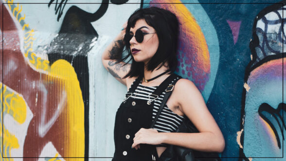 An alternative dressed girl with raven black hair and tattoos and wearing sunglasses, black overalls, and a striped crop top in front of an abstract wall mural