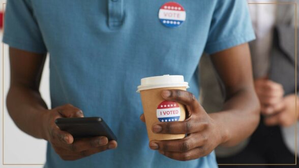 Man looking down at phone and holding a coffee to go cup with an "I Voted" sticker on it