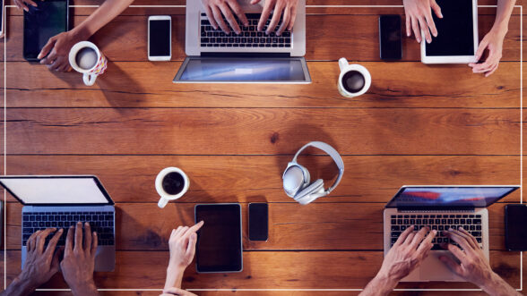 Image of a group of people sitting around a rectangular table with their laptops with coffee, headphones, and cell phones on the table.